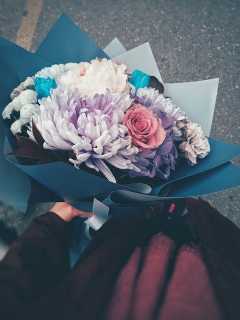 A bouquet of fresh flowers featuring a large, pale purple chrysanthemum, a single pink rose, white spray carnations, and bright blue accents, arranged with green foliage. The flowers are wrapped in el