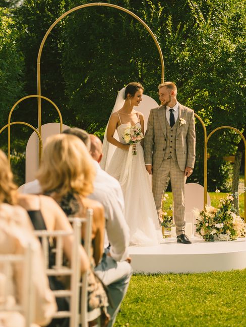 A bride and groom stand outdoors during their wedding ceremony, with natural light filtering through lush green trees in the background. The bride wears a classic white wedding gown with thin straps a