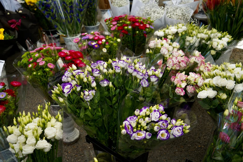 A vibrant display of fresh flowers arranged in bouquets and floral bunches, featuring a mix of white, pink, purple, and red blooms, with some roses, lisianthus, and miniature chrysanthemums. The flowe