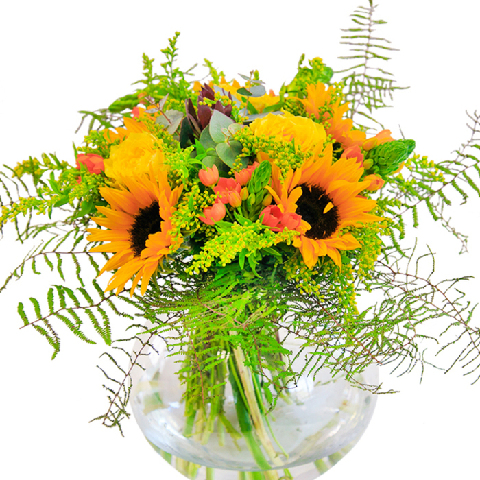 Drawing inspiration from the vibrant community of Cricklewood, this lush bouquet radiates the carefree energy of a sunlit day on Walm Lane. Sunflowers with bold, amber-yellow petals and rich brown centers seem to bask in invisible rays, creating a natural focal point that instantly warms any room. Nestled closely, yellow garden roses reveal countless layers that open slowly, reminiscent of morning blooms gently unfolding in local gardens. Threaded between the golden faces, deep green fern fronds drape in airy arches, their feathery texture suggesting a breeze drifting down Elm Grove. Clusters of glossy red Hypericum berries glisten like precious gems, adding both colour and a touch of playful elegance to the arrangement. The supporting foliage-ranging from ethereal solidago to slender flower spikes and soft eucalyptus-brings movement and a harmonious blend of green hues. All elements are gathered in a rounded, crystal-clear vase, where the submerged stems and shimmering water offer a fresh, modern presentation. The composition is both detailed and abundant, with a delicate interplay of textures: the velvety sunflowers, spiralled rose petals, and crisp greenery. A light scent, bright and inviting, lingers in the air, carrying silent memories of nearby allotment walks. Perfect for celebrations large or small-whether a joyful birthday, heartfelt anniversary, or simply to add cheer-this arrangement from Flowers Cricklewood delivers natural beauty and heartfelt artistry to every household in NW2.